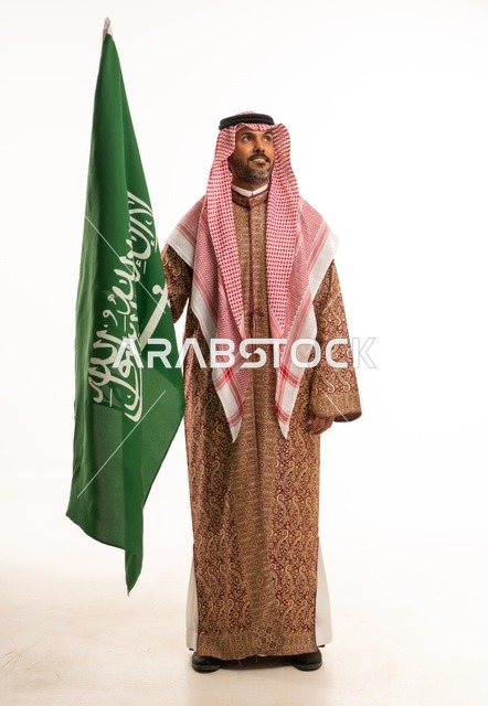 يوم العلم 11 مارس، ارتداء الازياء الشعبية في المناسبات الوطنية، اليوم الوطني السعودي 23 سبتمبر، الفخر والاعتزاز بالوطن، بورتريه لرجل عربي خليجي سعودي يرتدي الشماغ والدقلة يحمل بيده العلم السعودي، تأسيس المملكة السعودية 22 فبراير ، صورة الجسم بالكامل، خلفية بيضاء