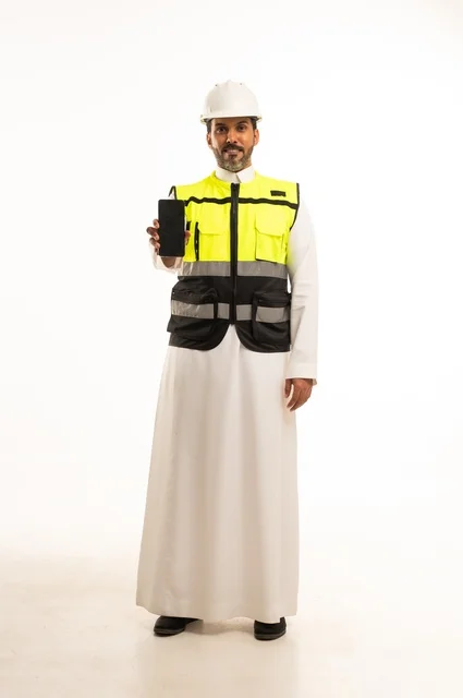 Display of a blank black mobile screen, working in the engineering sector, construction by the hands of the nation's sons, the concept of engineering and architectural constructions, a portrait of an Arab Gulf Saudi engineer wearing traditional attire and a special jacket and safety helmet standing and holding a mobile phone, full-body, white background.