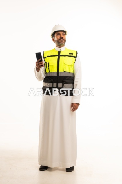 Working in the engineering sector, construction with the hands of the nation's sons, the concept of engineering and architectural constructions, a portrait of a Saudi Gulf Arab engineer wearing traditional attire and a special jacket and safety helmet, standing and holding a mobile phone, full-body, white background.