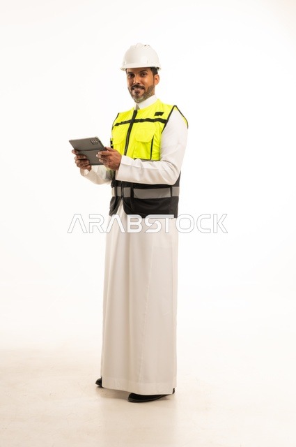 استخدام جهاز تقني حديث متطور في مجال الهندسة، العمل في القطاع الهندسي السعودي، مفهوم ادارة المشاريع الهندسية والانشاءات، مهنة ووظيفة شبابية، بورتريه لشاب مهندس عربي خليجي سعودي يرتدي الثوب التقليدي والسترة وخوذة الحماية يعمل على جهاز التابلت، خلفية بيضاء