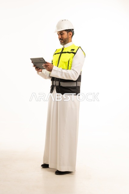 استخدام جهاز تقني حديث متطور في مجال الهندسة، العمل في القطاع الهندسي السعودي، مفهوم ادارة المشاريع الهندسية والانشاءات، مهنة ووظيفة شبابية، بورتريه لشاب مهندس عربي خليجي سعودي يرتدي الثوب التقليدي والسترة وخوذة الحماية يعمل على جهاز التابلت، خلفية بيضاء