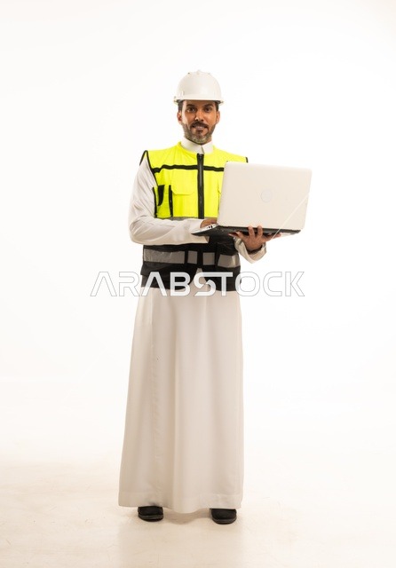 مفهوم ادارة المشاريع الهندسية والانشاءات، استخدام جهاز تقني حديث متطور في مجال الهندسة، العمل في القطاع الهندسي السعودي، مهنة ووظيفة شبابية، بورتريه لشاب مهندس عربي خليجي سعودي يرتدي الثوب التقليدي والسترة وخوذة الحماية يعمل على جهاز اللابتوب، خلفية بيضاء