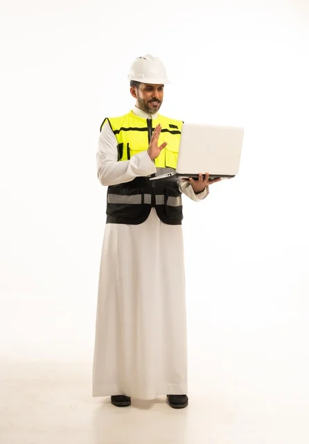 مفهوم ادارة المشاريع الهندسية والانشاءات، استخدام جهاز تقني حديث متطور في مجال الهندسة، العمل في القطاع الهندسي السعودي، مهنة ووظيفة شبابية، بورتريه لشاب مهندس عربي خليجي سعودي يرتدي الثوب التقليدي والسترة وخوذة الحماية يعمل على جهاز اللابتوب، خلفية بيضاء