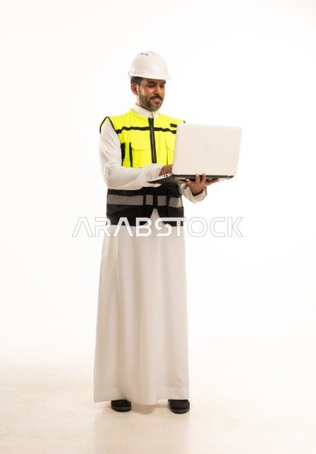 مفهوم ادارة المشاريع الهندسية والانشاءات، استخدام جهاز تقني حديث متطور في مجال الهندسة، العمل في القطاع الهندسي السعودي، مهنة ووظيفة شبابية، بورتريه لشاب مهندس عربي خليجي سعودي يرتدي الثوب التقليدي والسترة وخوذة الحماية يعمل على جهاز اللابتوب، خلفية بيضاء