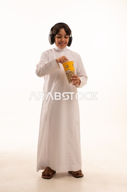 الاستمتاع بتناول سناك خفيف مع الاستماع إلى موسيقى، تناول الفوشار اللذيذ، بورتريه لفتى عربي خليجي سعودي يرتدي الزي التقليدي يحمل بيده علبة من الفشار الطازة ، الترفيه عن النفس وقضاء وقت ممتع، صورة الجسم بالطول الكامل، خلفية بيضاء