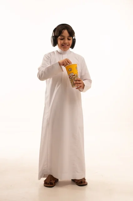 الاستمتاع بتناول سناك خفيف مع الاستماع إلى موسيقى، تناول الفوشار اللذيذ، بورتريه لفتى عربي خليجي سعودي يرتدي الزي التقليدي يحمل بيده علبة من الفشار الطازة ، الترفيه عن النفس وقضاء وقت ممتع، صورة الجسم بالطول الكامل، خلفية بيضاء