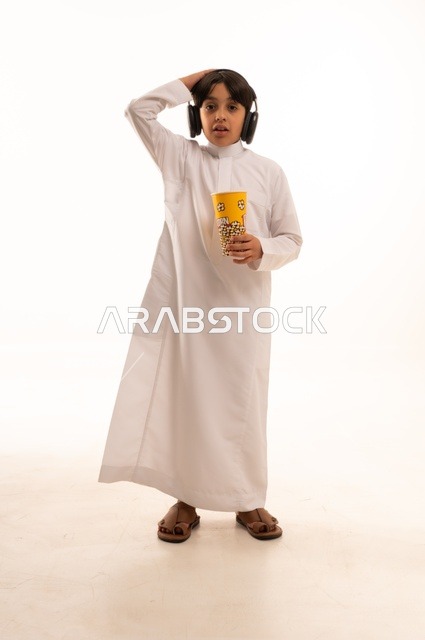 تعابير يد على الرأس تدل على الصدمة مع الاستماع إلى موسيقى، تناول الفوشار اللذيذ، بورتريه لفتى عربي خليجي سعودي يرتدي الزي التقليدي يحمل بيده علبة من الفشار الطازة ، الترفيه عن النفس وقضاء وقت ممتع، صورة الجسم بالطول الكامل، خلفية بيضاء