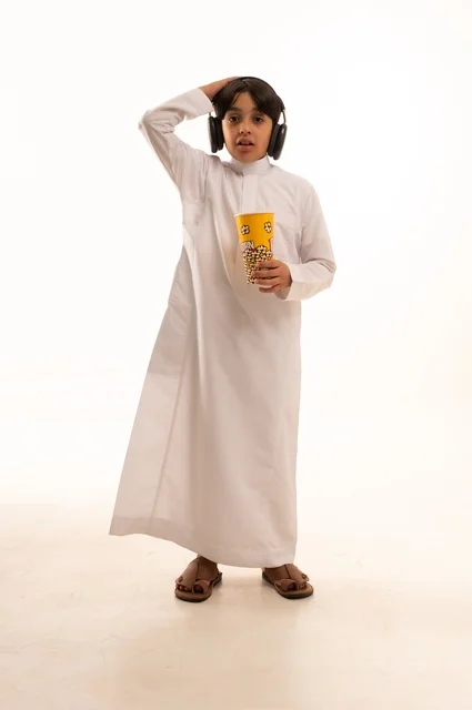 تعابير يد على الرأس تدل على الصدمة مع الاستماع إلى موسيقى، تناول الفوشار اللذيذ، بورتريه لفتى عربي خليجي سعودي يرتدي الزي التقليدي يحمل بيده علبة من الفشار الطازة ، الترفيه عن النفس وقضاء وقت ممتع، صورة الجسم بالطول الكامل، خلفية بيضاء