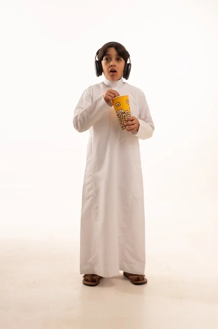 ايماءات الدهشة والحزن مع الاستماع إلى موسيقى، تناول الفوشار اللذيذ، بورتريه لفتى عربي خليجي سعودي يرتدي الزي التقليدي يحمل بيده علبة من الفشار الطازة ، الترفيه عن النفس وقضاء وقت ممتع، صورة الجسم بالطول الكامل، خلفية بيضاء