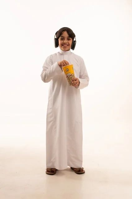 الاستماع إلى موسيقى مع ايمائات السرور والاستمتاع، تناول الفوشار اللذيذ، بورتريه لفتى عربي خليجي سعودي يرتدي الزي التقليدي يحمل بيده علبة من الفشار الطازة ، الترفيه عن النفس وقضاء وقت ممتع، صورة الجسم بالطول الكامل، خلفية بيضاء