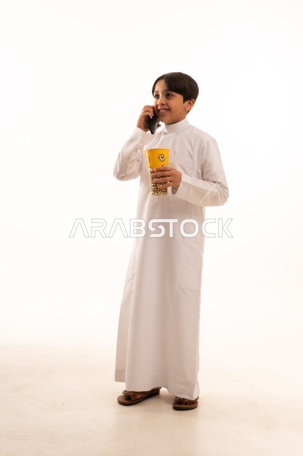 Making a phone call using a mobile device, enjoying delicious popcorn, a portrait of a young Arab Gulf Saudi boy wearing traditional attire holding a box of fresh popcorn in his hand, entertaining oneself and having a good time, full-body image, white background.