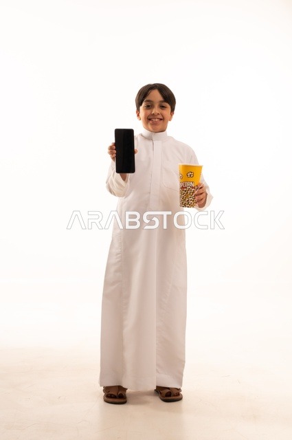 عرض خلفية شاشة سوداء على الهاتف المحمول، تناول الفوشار اللذيذ، بورتريه لفتى عربي خليجي سعودي يرتدي الزي التقليدي يحمل بيده علبة من الفشار الطازة ، الترفيه عن النفس وقضاء وقت ممتع، صورة الجسم بالطول الكامل، خلفية بيضاء