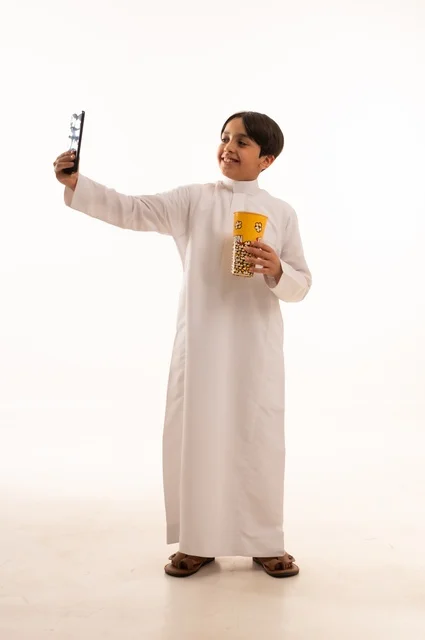 التقاط صورة سيلفي للأحتفاظ بالذكريات، تناول الفوشار اللذيذ، بورتريه لفتى عربي خليجي سعودي يرتدي الزي التقليدي يحمل بيده علبة من الفشار الطازة ، الترفيه عن النفس وقضاء وقت ممتع، صورة الجسم بالطول الكامل، خلفية بيضاء