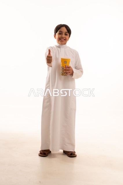 إشارة يد الأبهام للأعلى دليل علة الأعجاب، تناول الفوشار اللذيذ، بورتريه لفتى عربي خليجي سعودي يرتدي الزي التقليدي يحمل بيده علبة من الفشار الطازة ، الترفيه عن النفس وقضاء وقت ممتع، صورة الجسم بالطول الكامل، خلفية بيضاء