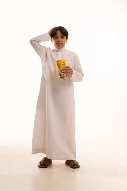 Enjoying delicious popcorn, a portrait of a young Arab Gulf Saudi boy wearing traditional attire, raising his hand up with gestures of surprise and astonishment, holding a box of fresh popcorn in his hand, entertaining himself and having a good time, full-body image, white background.
