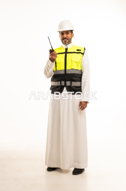 Giving specific work orders, the concept of engineering and architectural construction, using a wireless communication device at work, a portrait of an Arab Gulf engineer from Saudi Arabia wearing traditional attire with a jacket and safety helmet, full-body, white background.