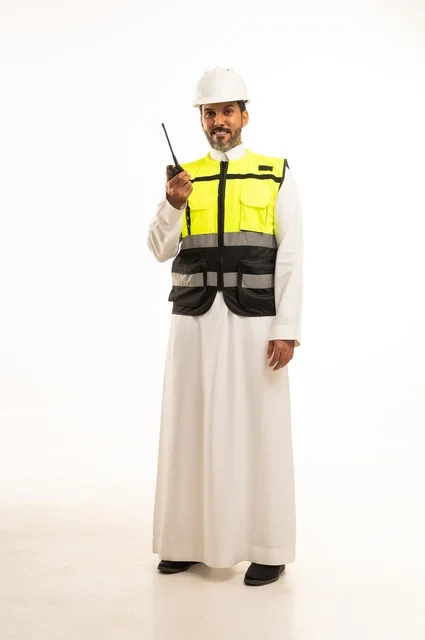 Giving specific work orders, the concept of engineering and architectural construction, using a wireless communication device at work, a portrait of an Arab Gulf engineer from Saudi Arabia wearing traditional attire with a jacket and safety helmet, full-body, white background.