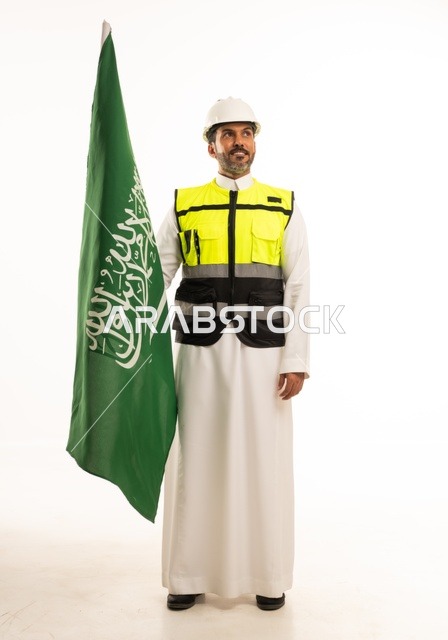التطور العمراني بسواعد أبناء الوطن ، الاحتفال بيوم العلم 11 مارس، ذكرى اليوم الوطني السعودي 8 ربيع الاول، بورتريه لمهندس عربي خليجي سعودي مبتسم يرتدي سترة العمل وخوذة الحماية البيضاء في اليوم الوطني ويحمل علم المملكة، الجسم بالطول الكامل، خلفية بيضاء