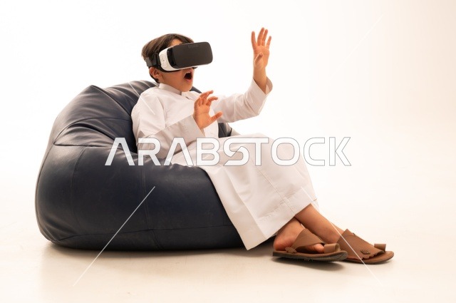 Interaction, fun, and entertainment from watching movies and virtual world games, a portrait of a happy Saudi Arabian Gulf child wearing unique and amazing 3D virtual reality glasses, pointing with his index finger at something he is watching, technologies and games, white background.
