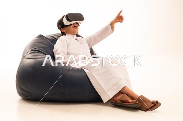 الإشارة باليد بأصبع السبابة لشئ ما يشاهده، بورتريه لطفل عربي خليجي سعودي سعيد يرتدي نظارة الواقع الإفتراضي ثلاثية الأبعاد الفريدة و العجيبة ،  التفاعل و المتعة و التسلية بمشاهدة الأفلام و ألعاب العالم الافتراضي ، تقنيات و ألعاب ، خلفية بيضاء