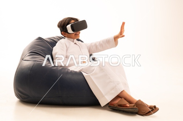 التفاعل و المتعة و التسلية بمشاهدة الأفلام و ألعاب العالم الافتراضي ، بورتريه لطفل عربي خليجي سعودي سعيد يرتدي نظارة الواقع الإفتراضي ثلاثية الأبعاد الفريدة و العجيبة ، الإشارة باليد بأصبع السبابة لشئ ما يشاهده، تقنيات و ألعاب ، خلفية بيضاء