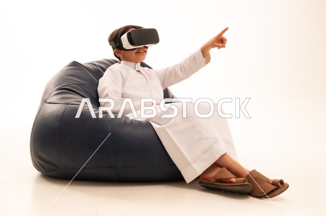 الإشارة باليد بأصبع السبابة لشئ ما يشاهده، بورتريه لطفل عربي خليجي سعودي سعيد يرتدي نظارة الواقع الإفتراضي ثلاثية الأبعاد الفريدة و العجيبة ،  التفاعل و المتعة و التسلية بمشاهدة الأفلام و ألعاب العالم الافتراضي ، تقنيات و ألعاب ، خلفية بيضاء