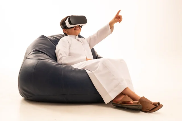 Pointing with the index finger at something he is watching, a portrait of a happy Saudi Arabian Gulf child wearing unique and amazing 3D virtual reality glasses, interacting and enjoying watching movies and playing virtual world games, technologies and games, white background.