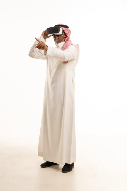 الاندماج والتفاعل مع العالم الخيالي، بورتريه لرجل عربي خليجي سعودي يرتدي الثوب والشماغ التقليدي ونظارات الواقع الافتراضي ثلاثية الأبعاد يحاول تجسيم شيء، دمج التقنيات والتكنولوجيا الحديثة مع المتعة والتسلية، الجسم بالطول الكامل، خلفية بيضاء