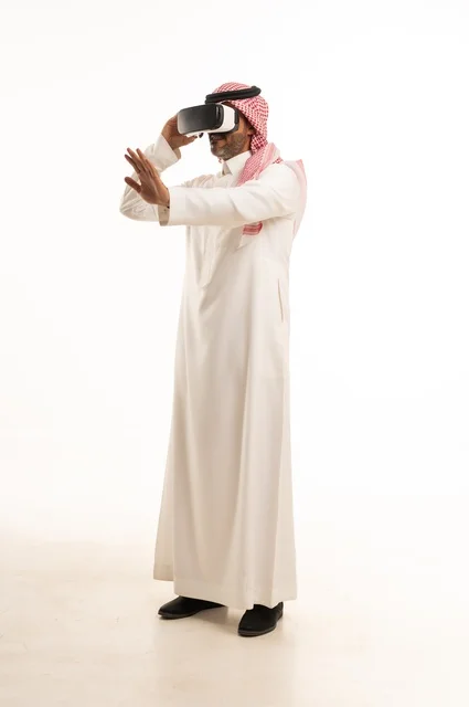 The integration and interaction with the imaginary world, a portrait of a Saudi Arabian Gulf man wearing traditional thobe and ghutrah, with 3D virtual reality glasses trying to embody something, merging modern technologies and technology with fun and entertainment, full-body, white background.