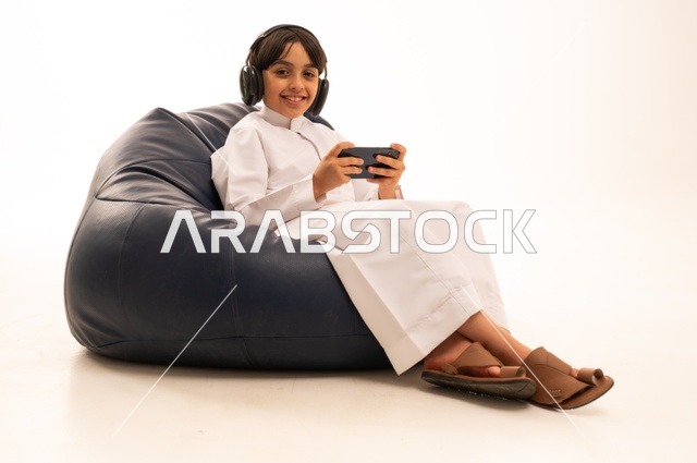 Looking at the camera with gestures of joy and enjoyment, using modern technologies for entertainment and amusement, a close-up portrait of an Arab Gulf Saudi boy sitting on a gray sofa wearing headphones and playing with a mobile phone, having fun playing, white background.