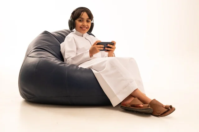 النظر إلى الكاميرا بإيماءات السرور والاستمتاع، استخدام التقنيات الحديثة للتسلية والترفيه، بورتريه مقرب لولد عربي خليجي سعودي يجلس على اريكة رمادية يضع سماعة رأس ويقوم باللعب باستخدام جهاز الهاتف محمول، قضاء اوقات ممتعة باللعب، خلفية بيضاء