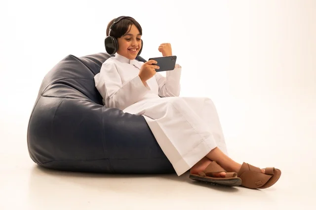 A fist gesture of victory and success, using modern technologies for entertainment and fun, a close-up portrait of an Arab Gulf Saudi boy sitting on a gray sofa wearing headphones and playing with a mobile phone, enjoying fun times playing, white background.