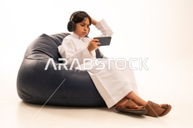 A hand on the head, gestures of regret and sadness, using modern technologies for entertainment and amusement, a close-up portrait of a Saudi Arabian Gulf boy sitting on a gray sofa wearing headphones and playing with a mobile phone, spending enjoyable times playing, white background.