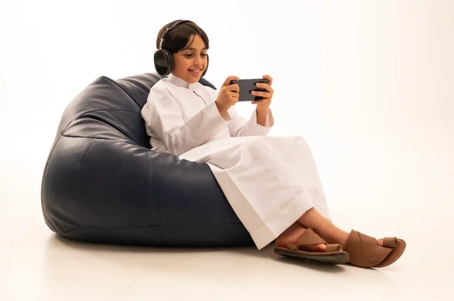 اللعب باستخدام جهاز الهاتف محمول، استخدام التقنيات الحديثة للتسلية والترفيه، بورتريه مقرب لولد عربي خليجي سعودي يجلس على اريكة رمادية يضع سماعة رأس ويقوم باللعب، قضاء اوقات ممتعة باللعب، خلفية بيضاء