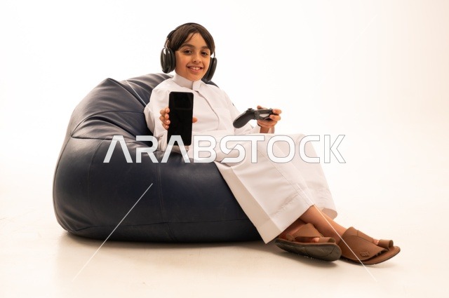 عرض خلفية شاشة سوداء على هاتف محمول، استخدام التقنيات الحديثة للتسلية والترفيه، بورتريه مقرب لولد عربي خليجي سعودي يجلس على اريكة رمادية يحمل عصا التحكم يقوم باللعب، قضاء اوقات ممتعة باللعب، خلفية بيضاء