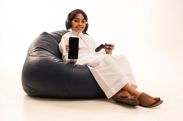 عرض خلفية شاشة سوداء على هاتف محمول، استخدام التقنيات الحديثة للتسلية والترفيه، بورتريه مقرب لولد عربي خليجي سعودي يجلس على اريكة رمادية يحمل عصا التحكم يقوم باللعب، قضاء اوقات ممتعة باللعب، خلفية بيضاء