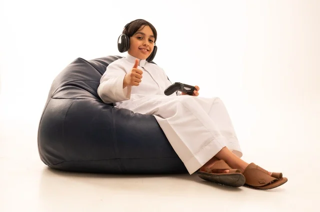 تعابير يد إشارة الأبهام للأعلى تدل على سير اللعب بطريقة مميزة، استخدام التقنيات الحديثة للتسلية والترفيه، بورتريه مقرب لولد عربي خليجي سعودي يجلس على اريكة رمادية يحمل عصا التحكم يقوم باللعب، قضاء اوقات ممتعة باللعب، خلفية بيضاء