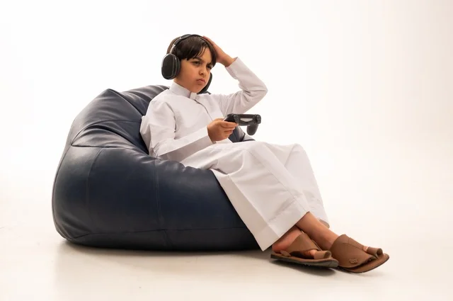 Hand expressions on the head indicate sadness and regret, the use of modern technologies for entertainment and amusement, a close-up portrait of an Arab Gulf Saudi boy sitting on a gray sofa holding a game controller while playing, spending enjoyable times playing, white background.
