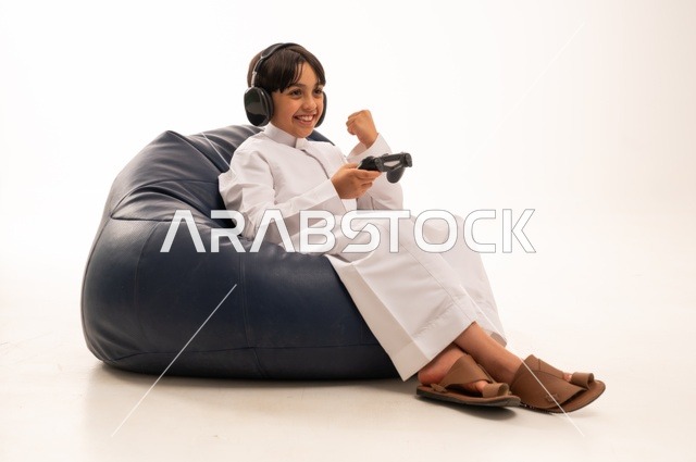 Hand gestures of a fist indicate victory and achieving goals, using modern technologies for entertainment and fun, a close-up portrait of a smiling Arab Gulf Saudi boy sitting on a gray sofa holding a game controller and playing, spending enjoyable times playing, white background.