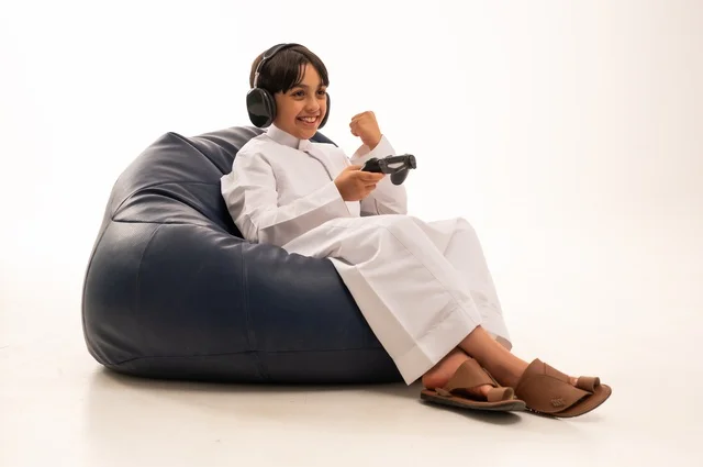 Hand gestures of a fist indicate victory and achieving goals, using modern technologies for entertainment and fun, a close-up portrait of a smiling Arab Gulf Saudi boy sitting on a gray sofa holding a game controller and playing, spending enjoyable times playing, white background.