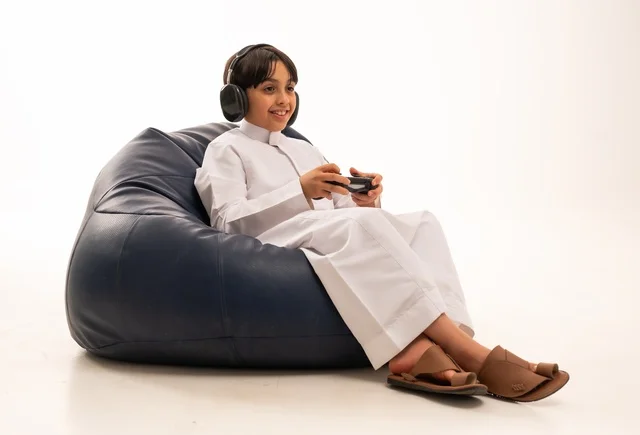 استخدام التقنيات الحديثة للتسلية والترفيه، بورتريه مقرب لولد عربي خليجي سعودي مبتسم يجلس على اريكة رمادية يحمل عصا التحكم يقوم باللعب، تعابير وايماءات الاندماج والتركيز، قضاء اوقات ممتعة باللعب، خلفية بيضاء