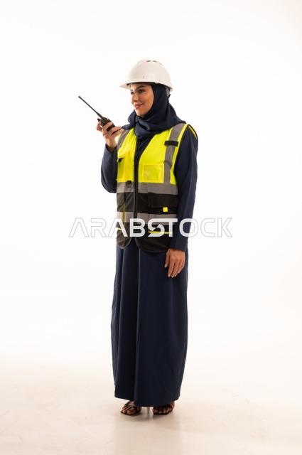 ااستخدام الجهاز اللاسلكي لمتابعة سير العمل، مهن ووظائف نسائية في القطاع الهندسي, دور الهندسة الإنشائية في تطور المجتمع السعودي, بورتريه لمهندسة عربية خليجية سعودية مسلمة محجبة ترتدي الخوذة البيضاء وسترة الحماية، الجسم بالطول الكامل، خلفية بيضاء