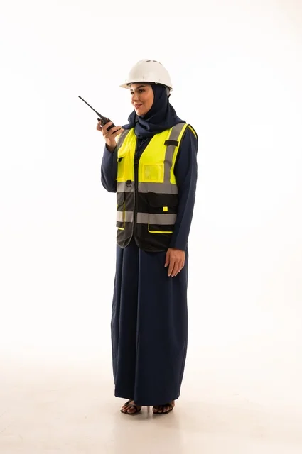 ااستخدام الجهاز اللاسلكي لمتابعة سير العمل، مهن ووظائف نسائية في القطاع الهندسي, دور الهندسة الإنشائية في تطور المجتمع السعودي, بورتريه لمهندسة عربية خليجية سعودية مسلمة محجبة ترتدي الخوذة البيضاء وسترة الحماية، الجسم بالطول الكامل، خلفية بيضاء