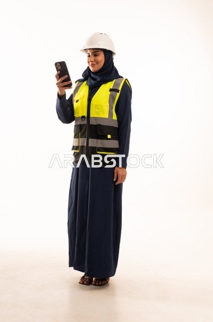 العمل على الهاتف المحمول والوقوف بظهر مشدود، مهن ووظائف نسائية في القطاع الهندسي, دور الهندسة الإنشائية في تطور المجتمع السعودي,  بورتريه لمهندسة عربية خليجية سعودية مسلمة محجبة ترتدي الخوذة البيضاء وسترة الحماية، الجسم بالطول الكامل، خلفية بيضاء