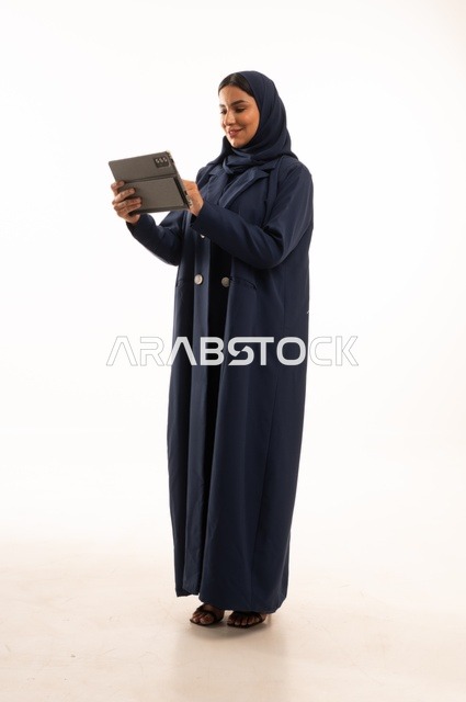 استخدام التقنية الحديثه المتطورة في الحياة العملية عبر الجهاز اللوحي، تعابير وايماءات الاندماج، بورتريه لشابة عربية خليجية سعودية مبتسمة ترتدي الحجاب والعباءة تقوم بالعمل على جهازالتابلت، صورة الجسم بالطول الكامل، خلفية بيضاء