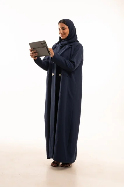 استخدام التقنية الحديثه المتطورة في الحياة العملية عبر الجهاز اللوحي، تعابير وايماءات الاندماج، بورتريه لشابة عربية خليجية سعودية مبتسمة ترتدي الحجاب والعباءة تقوم بالعمل على جهازالتابلت، صورة الجسم بالطول الكامل، خلفية بيضاء