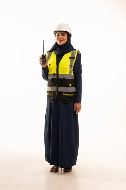 ااستخدام الجهاز اللاسلكي لمتابعة سير العمل، مهن ووظائف نسائية في القطاع الهندسي, دور الهندسة الإنشائية في تطور المجتمع السعودي, بورتريه لمهندسة عربية خليجية سعودية مسلمة محجبة ترتدي الخوذة البيضاء وسترة الحماية، الجسم بالطول الكامل، خلفية بيضاء