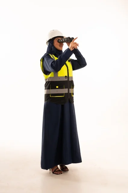 النظر من خلال منظار إلى شيء ما والوقوف بظهر مشدود، مهن ووظائف نسائية في القطاع الهندسي, دور الهندسة الإنشائية في تطور المجتمع السعودي,  بورتريه لمهندسة عربية خليجية سعودية مسلمة محجبة ترتدي الخوذة البيضاء وسترة الحماية، الجسم بالطول الكامل، خلفية بيضاء