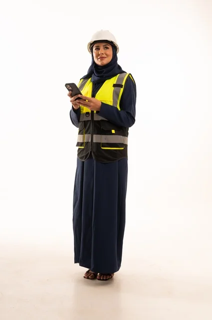 Working on a mobile phone and standing with a straight back, professions and jobs for women in the engineering sector, the role of structural engineering in the development of Saudi society, a portrait of a Gulf Arab Saudi Muslim engineer wearing a white helmet and safety vest, full-body, white background.