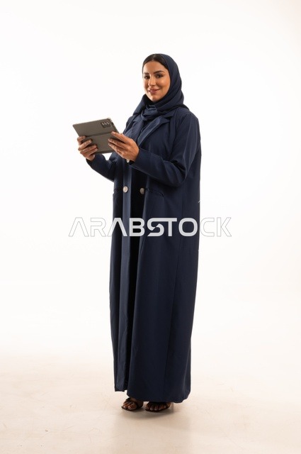استخدام التقنية الحديثه المتطورة في الحياة العملية عبر الجهاز اللوحي، تعابير وايماءات الاندماج، بورتريه لشابة عربية خليجية سعودية مبتسمة ترتدي الحجاب والعباءة تقوم بالعمل على جهازالتابلت، صورة الجسم بالطول الكامل، خلفية بيضاء